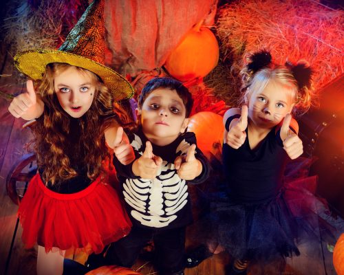 Three children dressed as a witch, a skeleton and a black cat celebrating halloween in a wooden barn with pumpkins. Halloween concept.