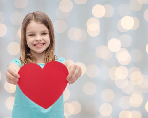 love, charity, holidays, children and people concept - smiling little girl with red heart over lights background